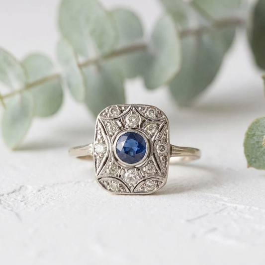 Art Deco Sapphire Diamond Ring featuring a 0.50 CT round cut deep blue natural sapphire in a square halo setting with round lab grown diamond accents and vintage inspired white metal detailing.