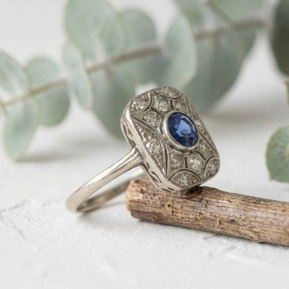 Art Deco Sapphire Diamond Ring featuring a 0.50 CT round cut deep blue natural sapphire in a square halo setting with round lab grown diamond accents and vintage inspired white metal detailing.