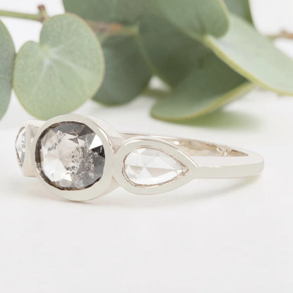 Oval cut salt and pepper diamond engagement ring featuring a 2.50 CT natural center stone with pear shaped side diamonds, set on a handcrafted knife edge band in a modern vintage fusion design.