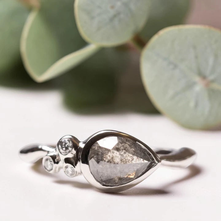 Pear Rose Cut Salt and Pepper Diamond Ring featuring a 1.50 CT natural salt and pepper diamond with organic inclusions, accented by two round cut diamond side stones in a handcrafted three stone design with a reverse tapered band and vintage inspired bridal elegance.