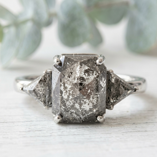 Rose Emerald Cut Salt and Pepper Diamond Ring featuring a 2.58 CT natural salt and pepper diamond with smoky galaxy-like inclusions, accented by triangle cut diamonds in a handcrafted, vintage-inspired, alternative engagement design with a straight 1.80 mm band.